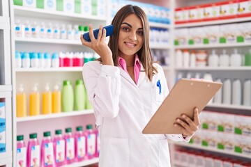 Young beautiful hispanic woman pharmacist listening voice message by smartphone reading document at pharmacy