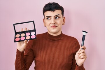Non binary person holding makeup brush and blush smiling looking to the side and staring away thinking.