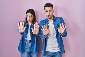 Young hispanic couple standing over pink background moving away hands palms showing refusal and...