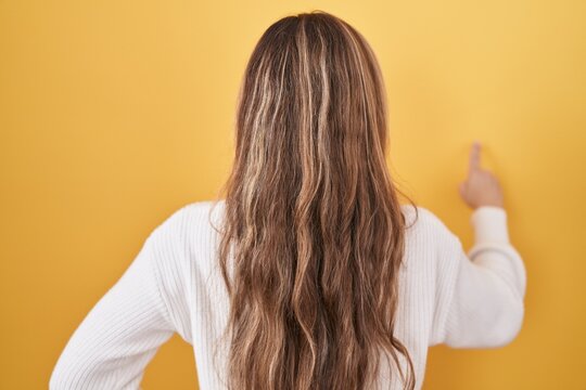 Young caucasian woman wearing white sweater over yellow background posing backwards pointing ahead with finger hand