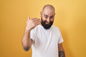 Young hispanic man with beard and tattoos standing over yellow background beckoning come here gesture with hand inviting welcoming happy and smiling