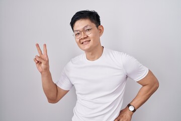 Young asian man standing over white background smiling looking to the camera showing fingers doing victory sign. number two.