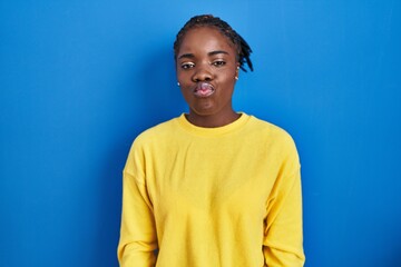 Beautiful black woman standing over blue background puffing cheeks with funny face. mouth inflated...