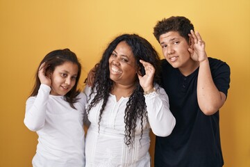 Family of mother, daughter and son standing over yellow background smiling with hand over ear listening an hearing to rumor or gossip. deafness concept.