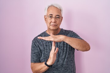 Middle age man with grey hair standing over pink background doing time out gesture with hands,...