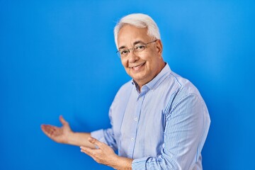 Hispanic senior man wearing glasses inviting to enter smiling natural with open hand