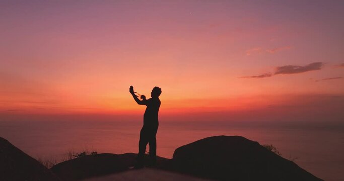 ..In a red sunset, a man takes a selfie on a cliff in bright red sky. .Nature video High quality footage. Meditation ocean and sky background. .Majestic sunset or sunrise cloudscape background..