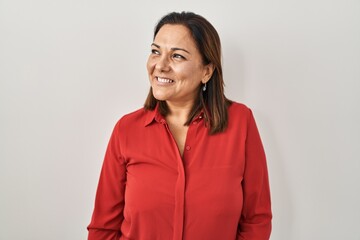 Hispanic mature woman standing over white background looking away to side with smile on face,...