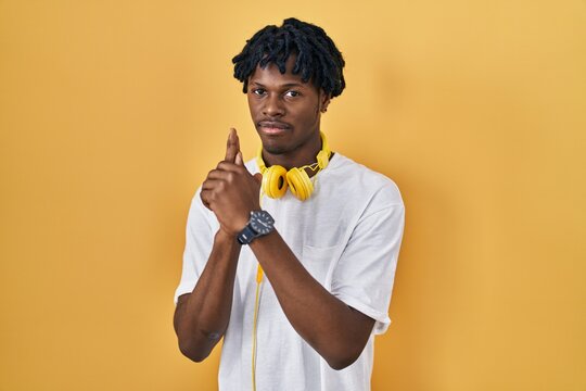 Young african man with dreadlocks standing over yellow background holding symbolic gun with hand gesture, playing killing shooting weapons, angry face