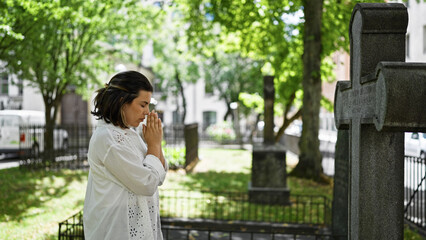 Beautiful young hispanic woman praying and mourning by tombstone at graveyard