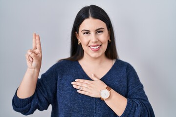 Young brunette woman standing over isolated background smiling swearing with hand on chest and...