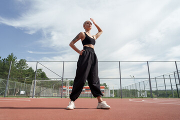 A photo of a blonde fitness model with a short haircut posing confidently on a red playground. She is wearing a black crop top and black baggy pants with white sneakers