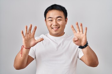 Young chinese man standing over white background showing and pointing up with fingers number nine...