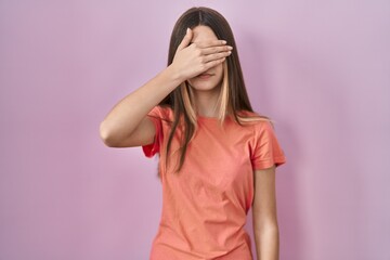 Teenager girl standing over pink background covering eyes with hand, looking serious and sad. sightless, hiding and rejection concept