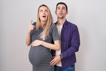 Young couple expecting a baby standing over white background amazed and surprised looking up and pointing with fingers and raised arms.