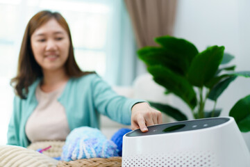 Happy Asian young woman turning on high efficiency air purifier while staying and relaxing in the living room.  - obrazy, fototapety, plakaty