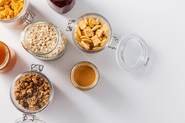 food storage and eating concept - close up of jars with oat, corn flakes, granola, cookies and spreads on white background
