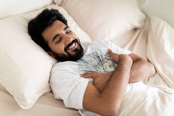 Smiling young indian man keeping dollar cash while sleeping in bed