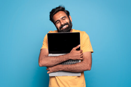Excited indian man hugging laptop with black blank screen, holding it tight near chest and smiling, copy space
