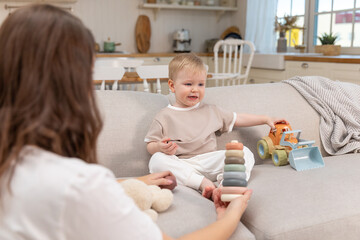 Happy family at home. Mother and baby boy playing with toys in couch at home indoors. Little toddler child and babysitter nanny having fun together. Young woman mom kid son rest in living room