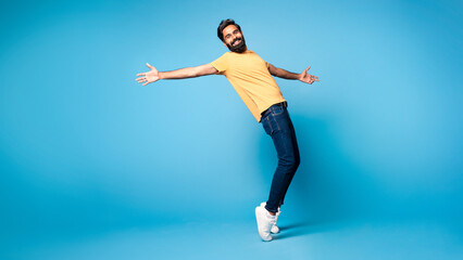 Positive funny middle aged indian man fooling on blue studio background, standing on toes, full length, panorama