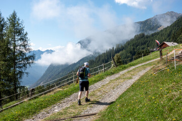 trekking man walking up gravel road on forest covered mountain to a malga, mountain hut, with alpine restaurant in Italian dolomites with low clouds 