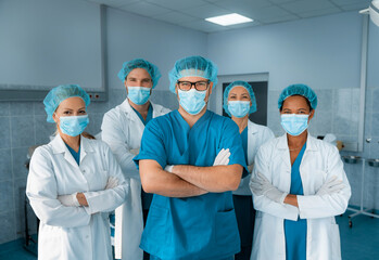 Group of medical professionals standing and looking at camera in the hospital operating room as they pose together for a portrait, dressed professionally in scrubs and lab coats wearing medical masks.