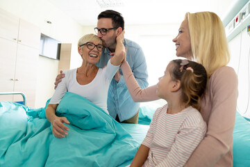 Cheerful smiling family visiting adult elder woman in hospital while son kisses her and daughter...