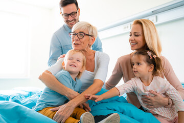 Joyful family with son and daughter visiting grandmother in hospital while hugging and talking to...