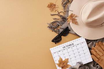 Capturing the spirit of autumn in a successful style. Top view shot of calendar, woolen hat, grey scarf, sunglasses, gloves, dry maple leaves on pastel brown background with space for advert or text