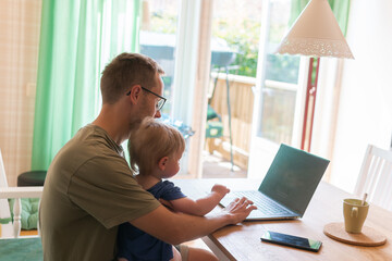A man in glasses sitting with little son at the computer indoors in a private house filled with sunlight. Freelancer, workation, work at home. Parenting, fatherhood. 