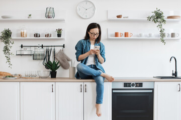 Lady using smartphone while sitting on countertop in kitchen