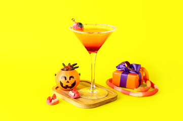Glass of tasty Margarita cocktail for Halloween, treats and gift box on yellow background
