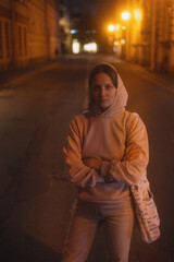 Fototapeta na wymiar young thoughtful woman stands on road in beige hoodie. walk in evening in light of lanterns, portrait of female alone. freedom loneliness of modern generation. Travel and rest alone, mental health