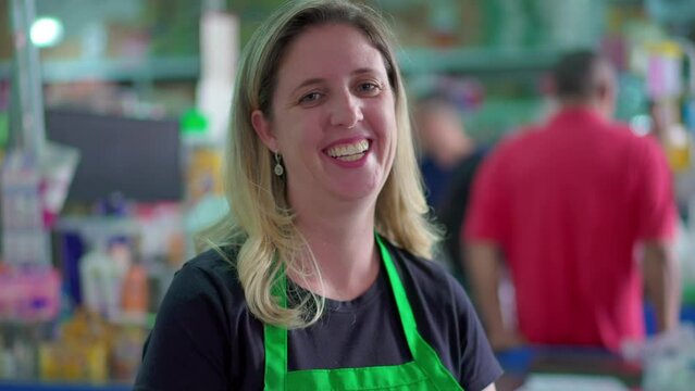 Cheerful Supermarket Employee Smiling at Camera, Shoppers in Background at Checkout