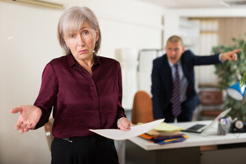Upset senior woman office worker holding documents in hand while her boss shouting at her in...