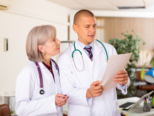 Two professional doctors standing in office, reading medical card of patient and discussing diagnosis