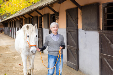 Mature woman jockey leads white horse by the bridle on the street along the stable