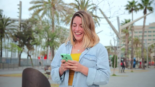 Slow motion fotage of one young adult woman walking and having fun using a cellphone social media application. Blonde feamale texting and sending funny messages on her smartphone. Lady holding phone