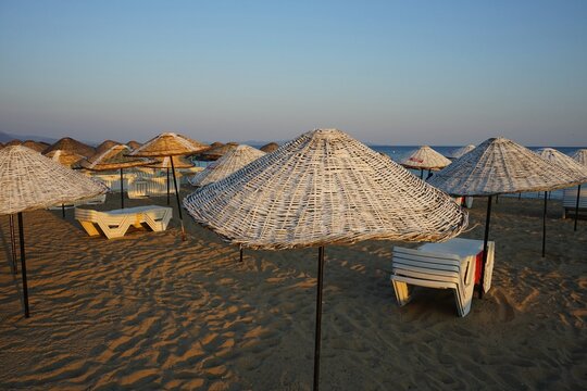 Weiße Liegen aus Kunststoff und Sonnenschirme aus Korbgeflecht vor im warmen Licht der untergehenden Abendsonne im Sommer am Strand von Sarimsakli bei Ayvalik in der Provinz Balikesir in der Türkei