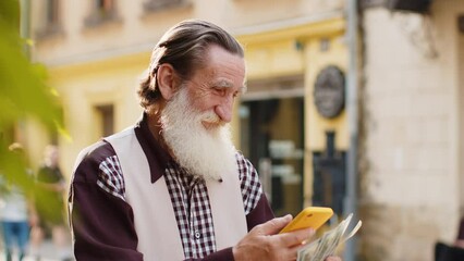 Happy rich senior old man counting holding money dollar cash, use smartphone calculator app, plans to order gifts, food delivery online, booking hotel room. Elderly grandfather tourist in city street - Powered by Adobe