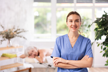 Successful confident young female massage therapist in blue scrub standing in procedure room of...