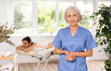 Obraz premium Successful confident mature female massage therapist standing in procedure room of modern wellness center, looking at camera with smile..