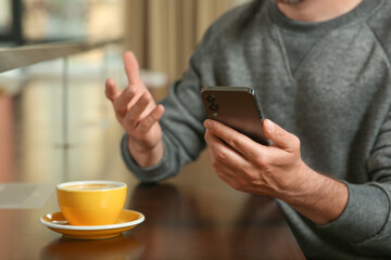 Handsome man with smartphone and cup of drink at table, closeup