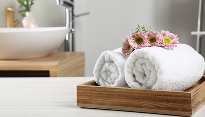 Wooden tray with stacked bath towels and beautiful flowers on white table in bathroom. Space for text