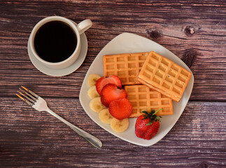 A cup of black coffee on a saucer, a fork and a plate with fresh Viennese waffles with strawberries and ripe banana slices on a wooden table.