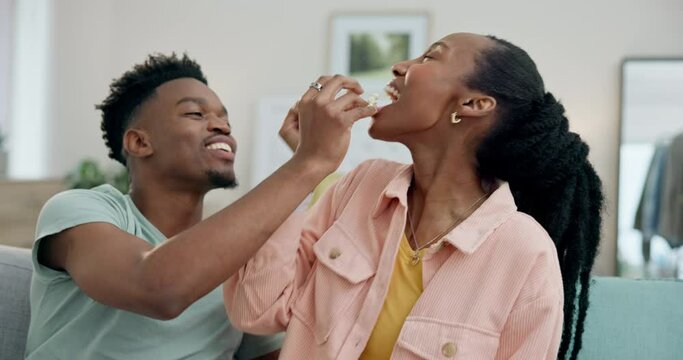 Black couple on sofa, feeding popcorn and love on date night together in living room in home. Relax, African man and happy woman eating snacks on couch with romance, fun and romance in apartment.