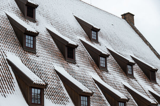 Snow falling on the roofs of the city. Snow-covered brown metal tiles roof of European house with windows. Rooftop covered in fresh snow. Winter scene
