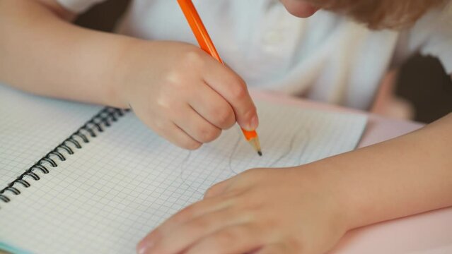 Schoolchild draws abstract picture with graphite pencil in checkered exercise book at desk closeup. Toddler boy does creative task at art lesson