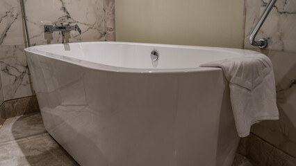 Bathroom interior. A white oval acrylic bathtub stands against a beige tiled wall. The towel is thrown over the edge. Shiny metal taps, valves, handrails.